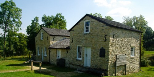 Historic stone building with a sloped roof, surrounded by trees and grass under a clear blue sky.
