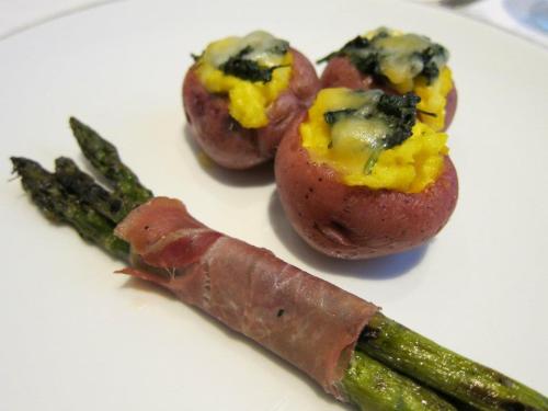 Three stuffed red potatoes topped with cheese and a side of asparagus wrapped in prosciutto on a white plate.