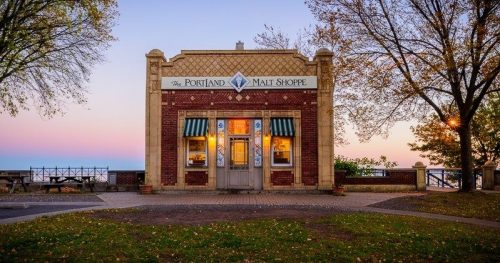 A charming brick shop with a sign reading "The Portland Malt Shoppe," surrounded by trees at sunset.