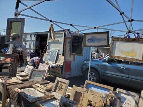 A market stall displaying framed artwork and photographs under a canopy, with a car parked nearby.