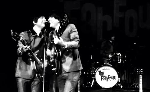 Two musicians in matching suits play guitars and sing closely together on stage, with a drummer in the background.