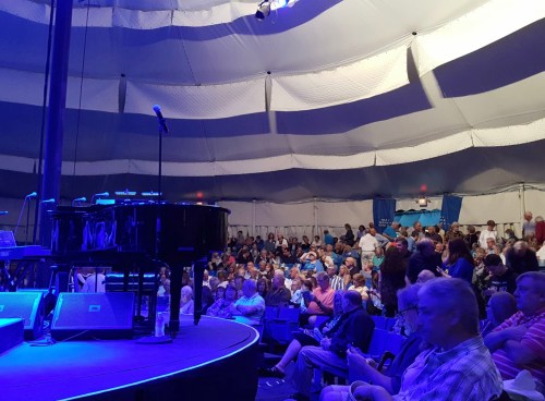 A crowded tent with a grand piano on stage, illuminated by blue lights, as an audience eagerly awaits a performance.