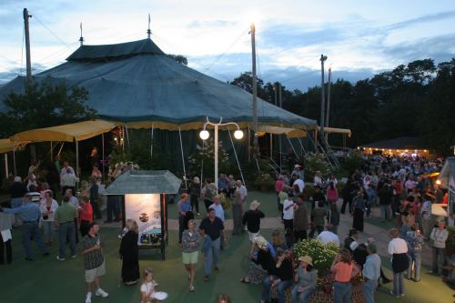 A bustling outdoor scene with a large circus tent, crowds of people, and evening sky in the background.