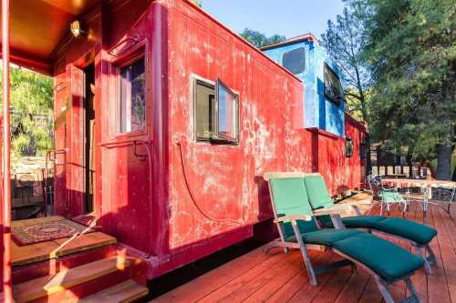 A colorful red and blue train caboose converted into a cozy space, with green chairs on a wooden deck.