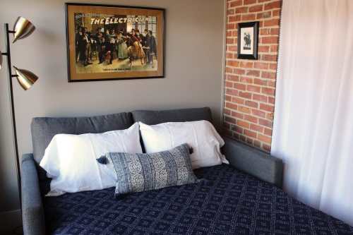 Cozy bedroom with a gray sofa bed, decorative pillows, and a vintage poster on the wall, featuring exposed brick.