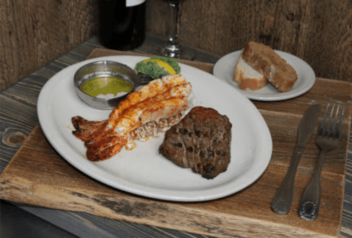 A plate with grilled lobster tail, steak, broccoli, and a side of butter, accompanied by a slice of bread.