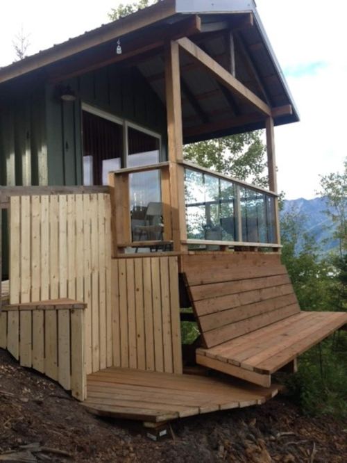 A wooden cabin with a deck and glass railing, set on a hillside surrounded by trees and mountains in the background.