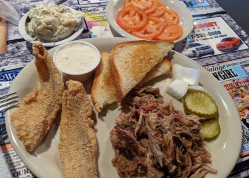 A plate with fried fish, pulled pork, toast, pickles, onions, and a side of sliced tomatoes and coleslaw.