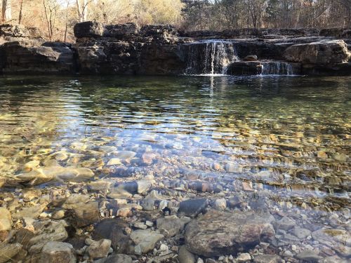 A serene natural scene featuring clear water over pebbles, with a small waterfall and rocky surroundings.