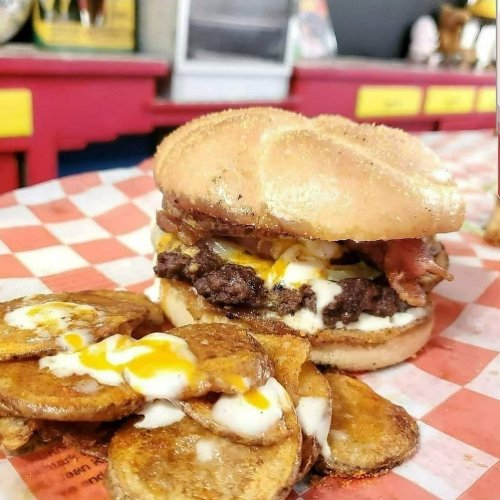 A hearty burger with bacon, cheese, and a toasted bun, served with crispy potato slices on a checkered plate.