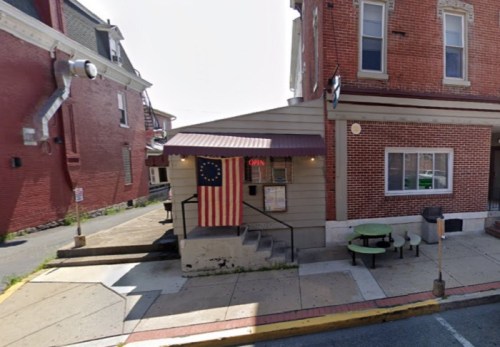 A small building with a flag, featuring an "OPEN" sign, located on a street with red brick buildings.
