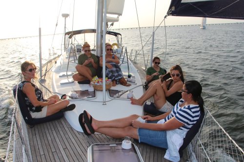 A group of six women relax on a sailboat, enjoying the water and sunshine. Snacks and drinks are nearby.