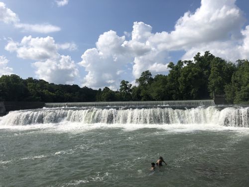 A waterfall cascades over a dam into a river, with two people swimming nearby under a blue sky with fluffy clouds.