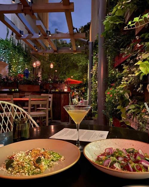 A cozy outdoor dining scene with two plates of food and a cocktail, surrounded by greenery and soft lighting.