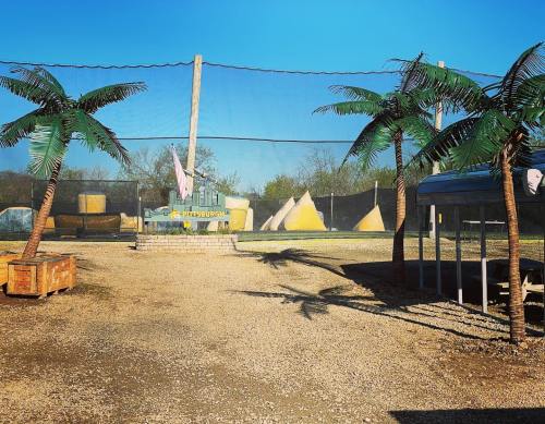A paintball field with palm trees, netting, and colorful obstacles under a clear blue sky.
