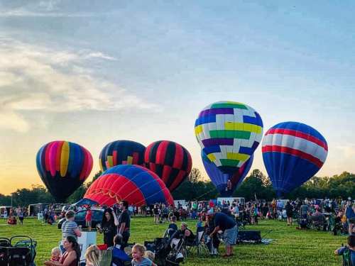 Colorful hot air balloons rise above a crowd gathered on a grassy field during a festival at sunset.