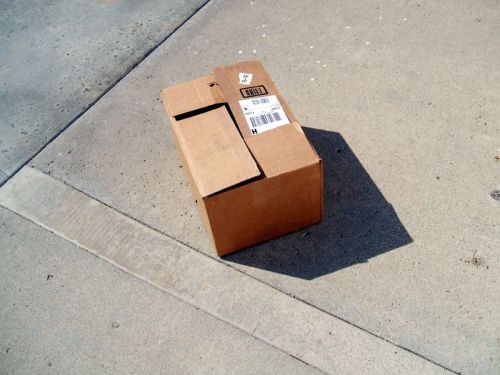 A cardboard box with a shipping label sits on a concrete surface, casting a shadow.