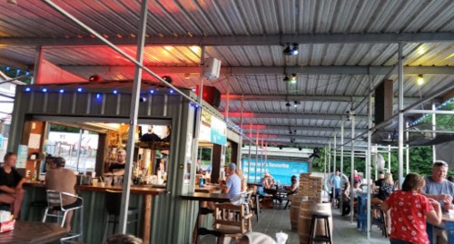 A lively outdoor bar scene with patrons seated at tables and a bar, under colorful lights and a metal roof.