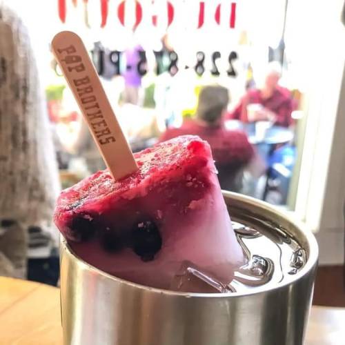 A colorful popsicle with berries sits in a drink, with people blurred in the background of a café.