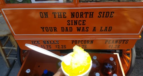 A hand holding a yellow snow cone in front of an orange cart with signs for ice balls, popcorn, and peanuts.