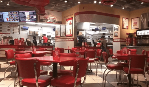 Interior of a fast-food restaurant with red tables and chairs, a counter, and staff preparing food in the background.