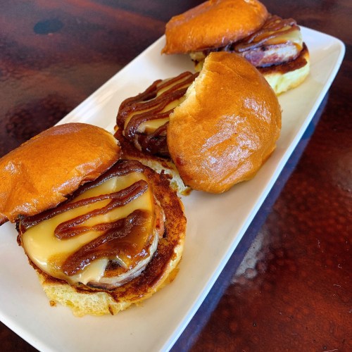 Three savory sliders with melted cheese and a drizzle of sauce on soft buns, served on a white plate.