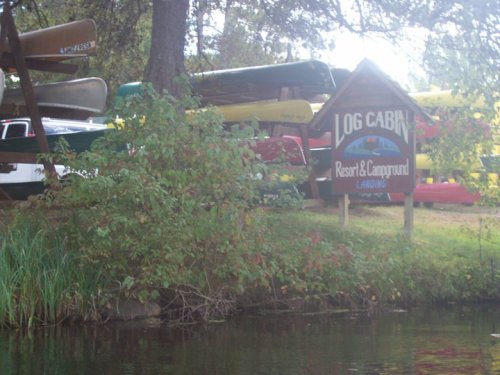 A sign for Log Cabin Resort & Campground, surrounded by canoes and lush greenery near a calm waterway.