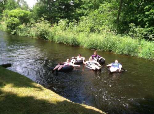 A group of five people relax on inner tubes in a calm, green-lined river on a sunny day.