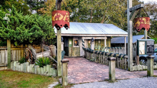 A rustic building with a metal roof, surrounded by greenery and wooden fences, featuring signs and festive decorations.