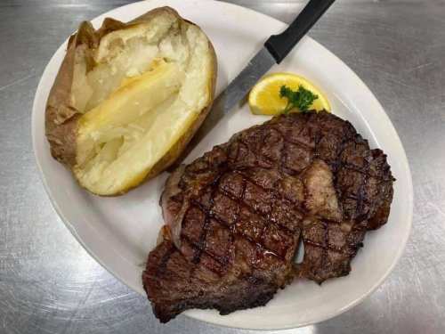 A grilled steak with a baked potato and a lemon wedge, garnished with parsley on a white plate.