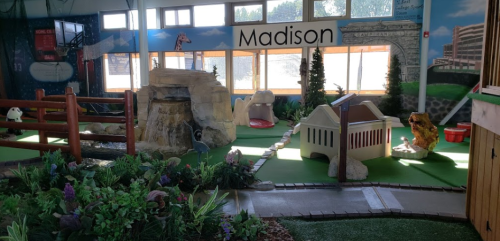 Indoor play area featuring a mini-golf course, rock formations, greenery, and themed decorations in a bright space.