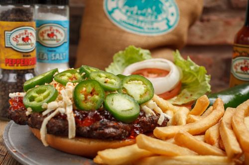 A close-up of a burger topped with jalapeños and cheese, served with fries and condiments in the background.