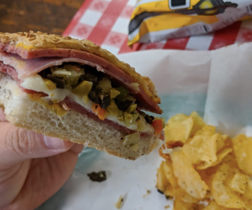 A hand holding a sandwich filled with meats, pickles, and vegetables, alongside a serving of potato chips.