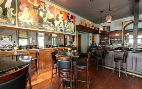A cozy bar interior featuring wooden furniture, colorful artwork on the walls, and a well-stocked bar area.
