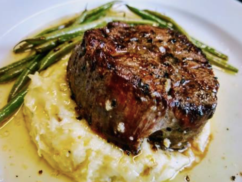 A perfectly cooked steak on a bed of creamy mashed potatoes, garnished with green beans and drizzled with sauce.