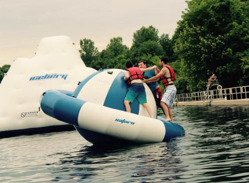 Three people in life jackets play on a large inflatable water toy in a lake, surrounded by greenery.