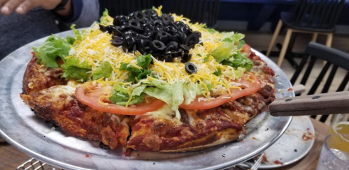 A colorful pizza topped with lettuce, tomatoes, black olives, and shredded cheese on a metal serving tray.
