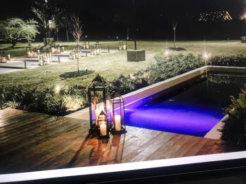 A serene outdoor space at night, featuring a pool with purple lighting and lanterns surrounded by greenery and candles.