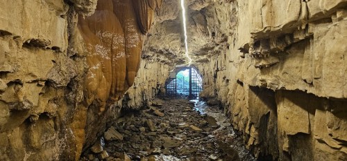 A dimly lit cave with rocky walls, a muddy floor, and a barred exit illuminated by a narrow beam of light.