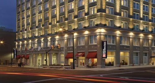 A well-lit hotel building at dusk, featuring large windows and red awnings, with street traffic in the foreground.