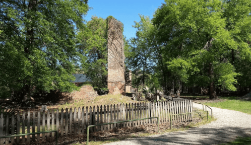Ruins of a brick chimney surrounded by trees and a wooden fence, with a clear blue sky above.