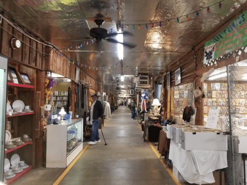 A long, narrow aisle in a vintage market with various booths, displays, and a person browsing. Colorful lights hang above.