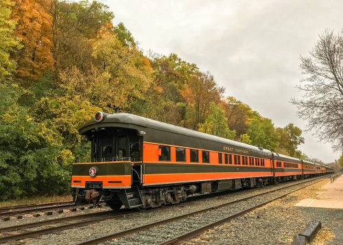 A vintage train with orange and black cars parked on tracks, surrounded by colorful autumn trees.