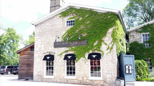 A stone building with ivy, featuring a sign that reads "Anvil Pub & Grille" and large windows.