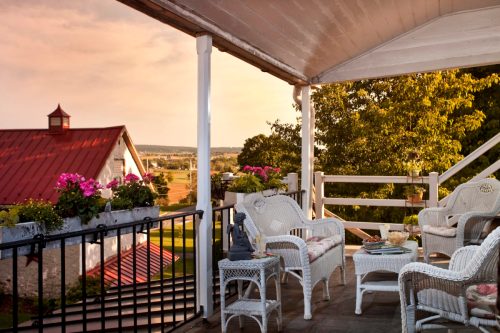 A cozy porch with white wicker furniture and potted flowers, overlooking a scenic landscape and a red-roofed barn.