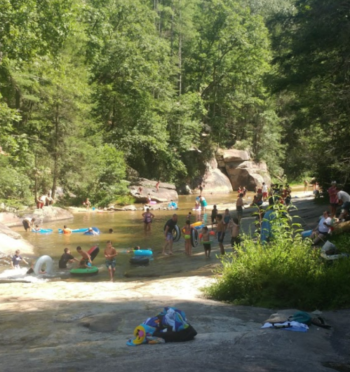 A busy river scene with people swimming, floating on tubes, and relaxing on rocks surrounded by lush greenery.
