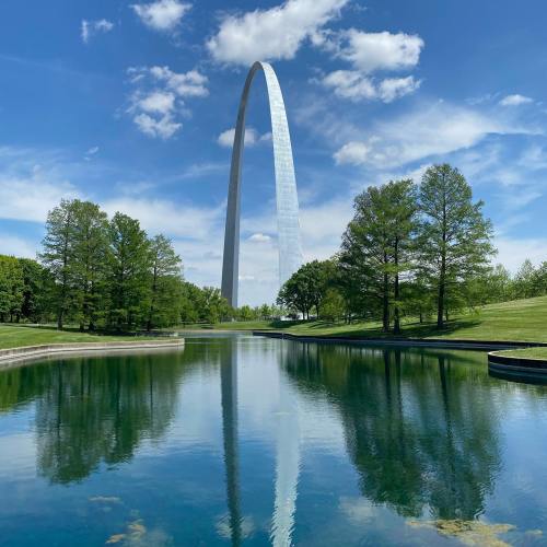 The Gateway Arch rises above a serene pond, surrounded by lush greenery and a bright blue sky with fluffy clouds.
