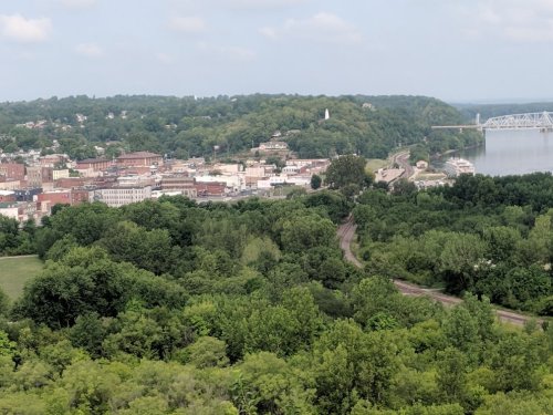 A scenic view of a town nestled among green hills, with a river and bridge in the background.