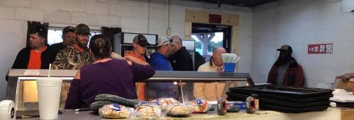 A group of people in a casual setting, waiting in line at a food counter with snacks and drinks visible.