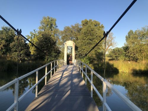 A suspension bridge over a calm river, surrounded by trees and greenery under a clear blue sky.